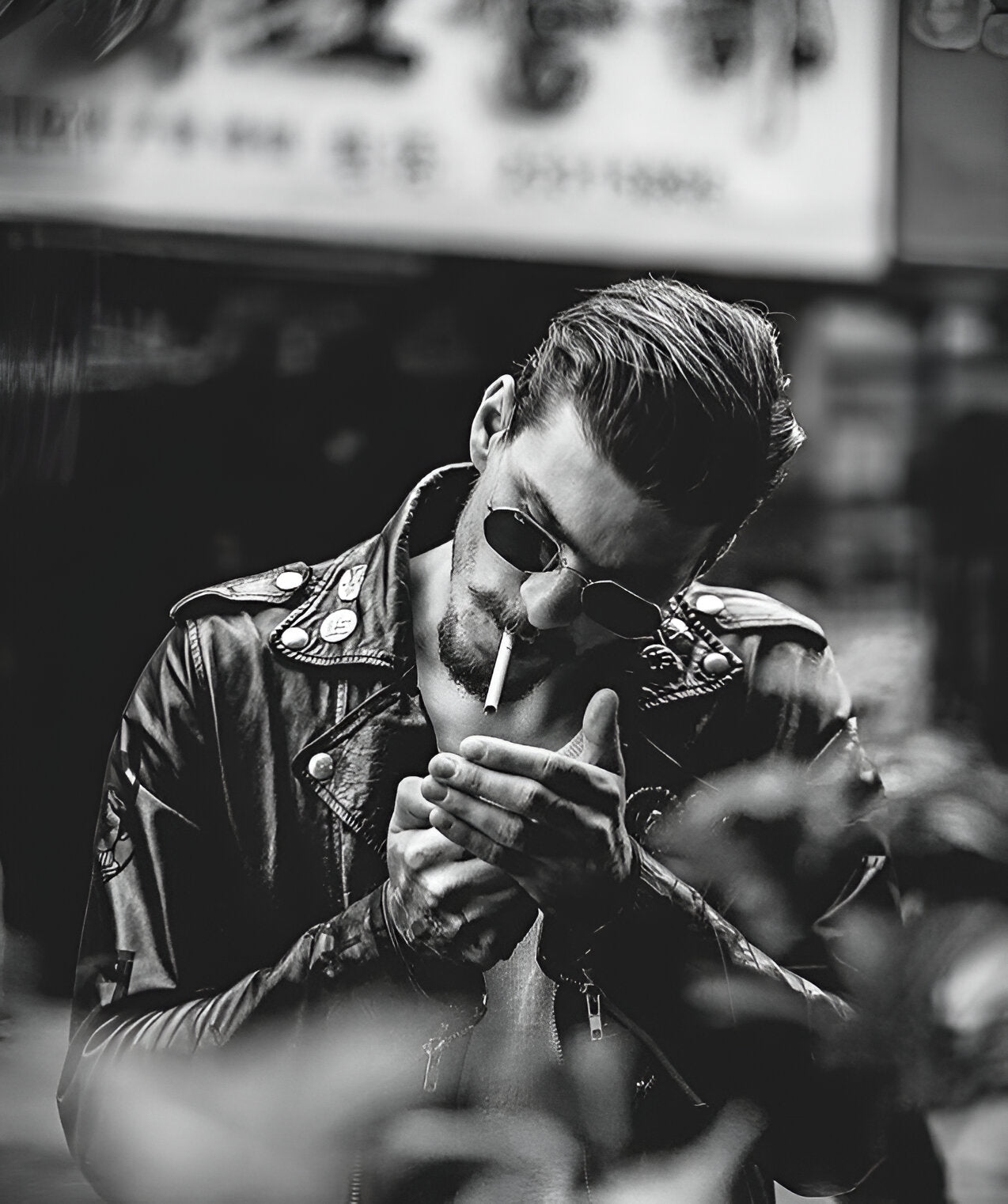 Hombre con actitud rebelde usando chamarra de piel negra con estoperoles, lentes de sol y encendiendo un cigarrillo, fotografía en blanco y negro estilo vintage